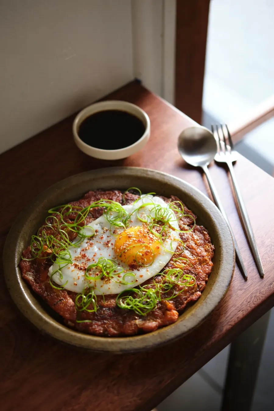 A kimchi pancake under a fried egg and a tangle of shredded spring onion in a stoneware dish, a cup of dipping sauce, a spoon and a fork beside it on a timber ledge by the window