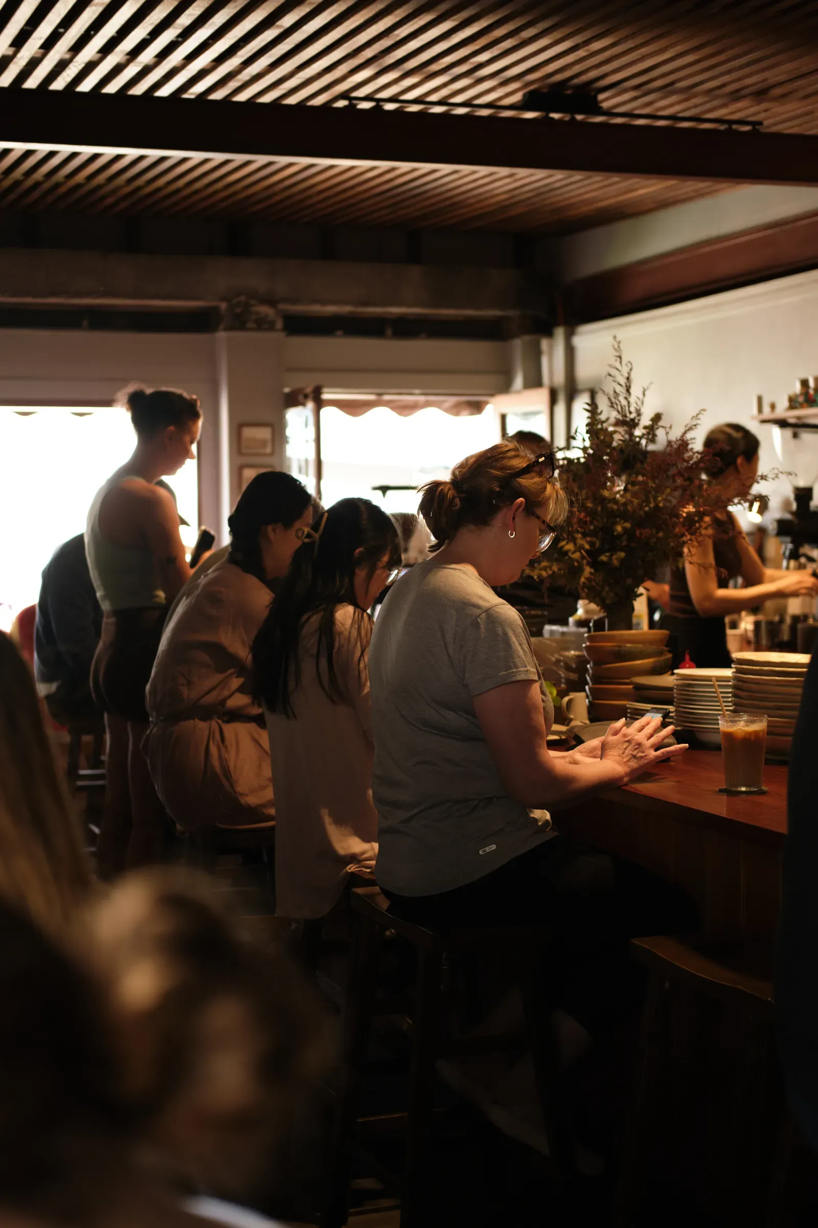 The bar by day, people on stools along the timber counter and one standing by the bright front windows, a staff member at the coffee machine behind