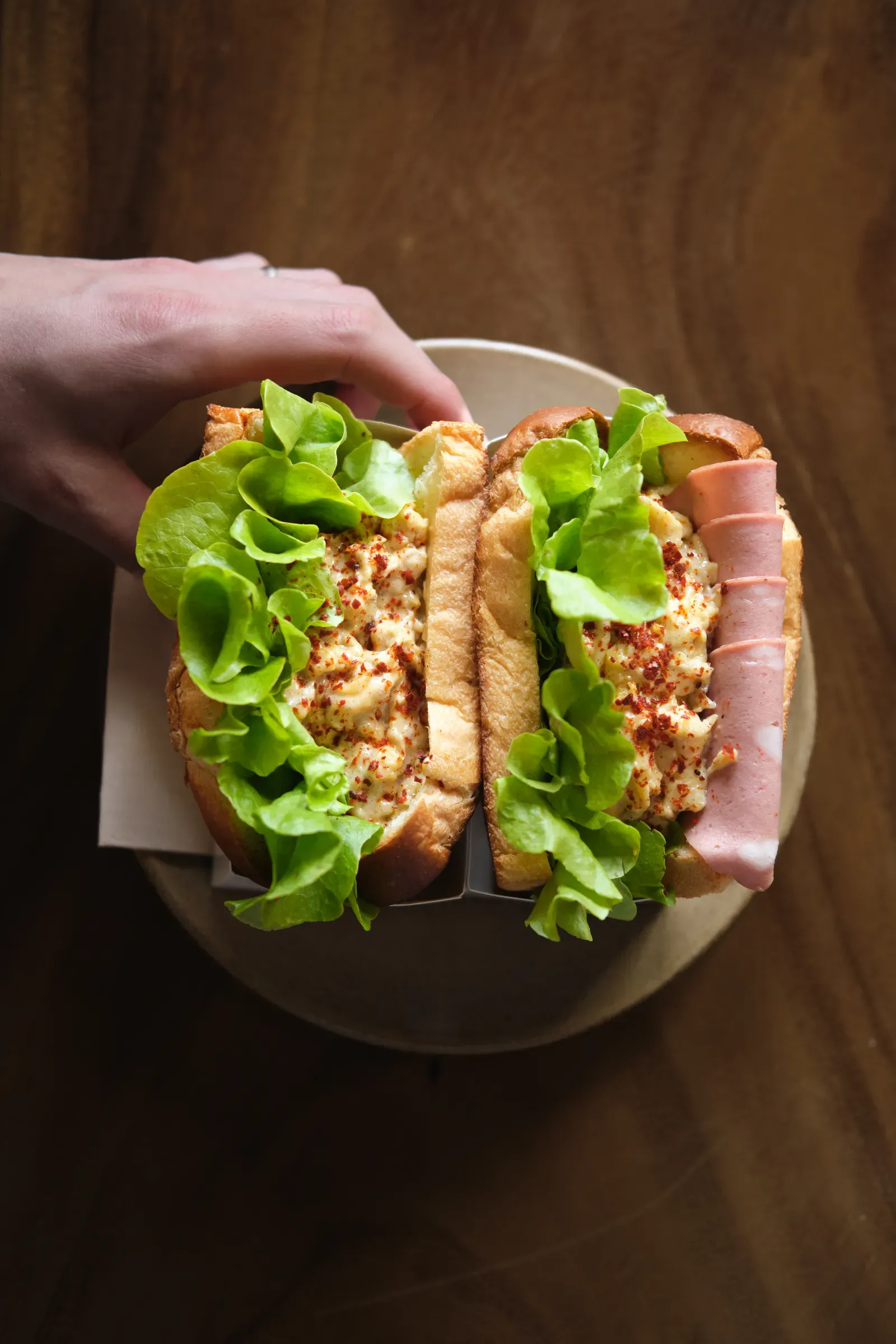 Two milk-bread sandwiches of scrambled egg, chilli flakes and butter lettuce, one with folded mortadella, held in a hand over a plate, seen from above