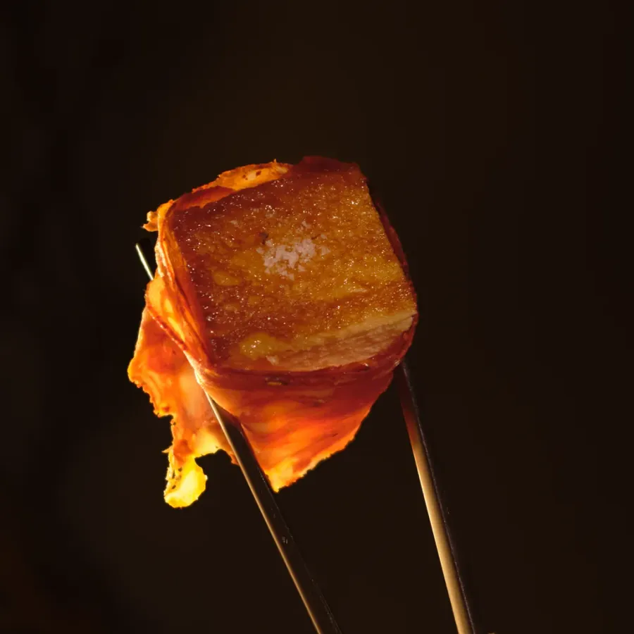 A cube of glossy pork held up in silver chopsticks, lit from behind against a dark room