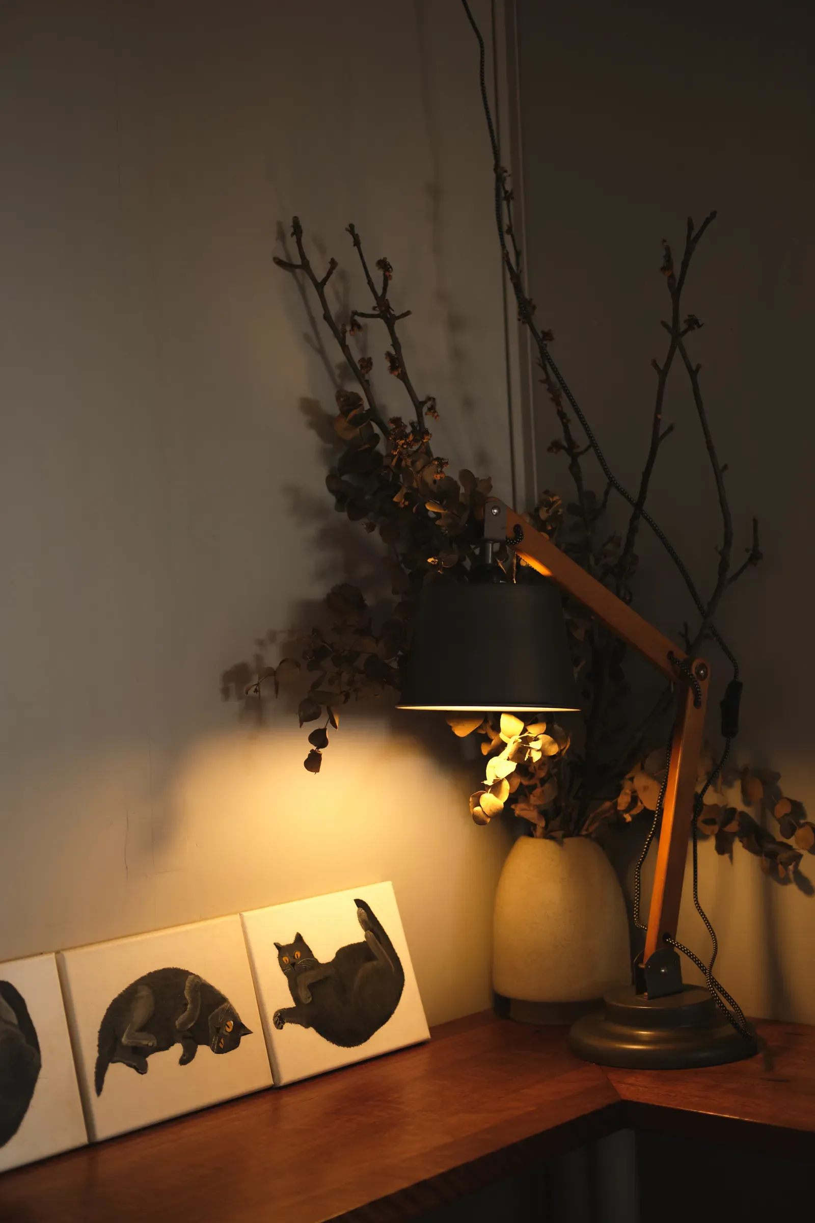 A desk lamp lighting a corner of the timber bench in the evening: dried branches and leaves in a cream vase, and small painted canvases of a dark grey cat leaning on the wall