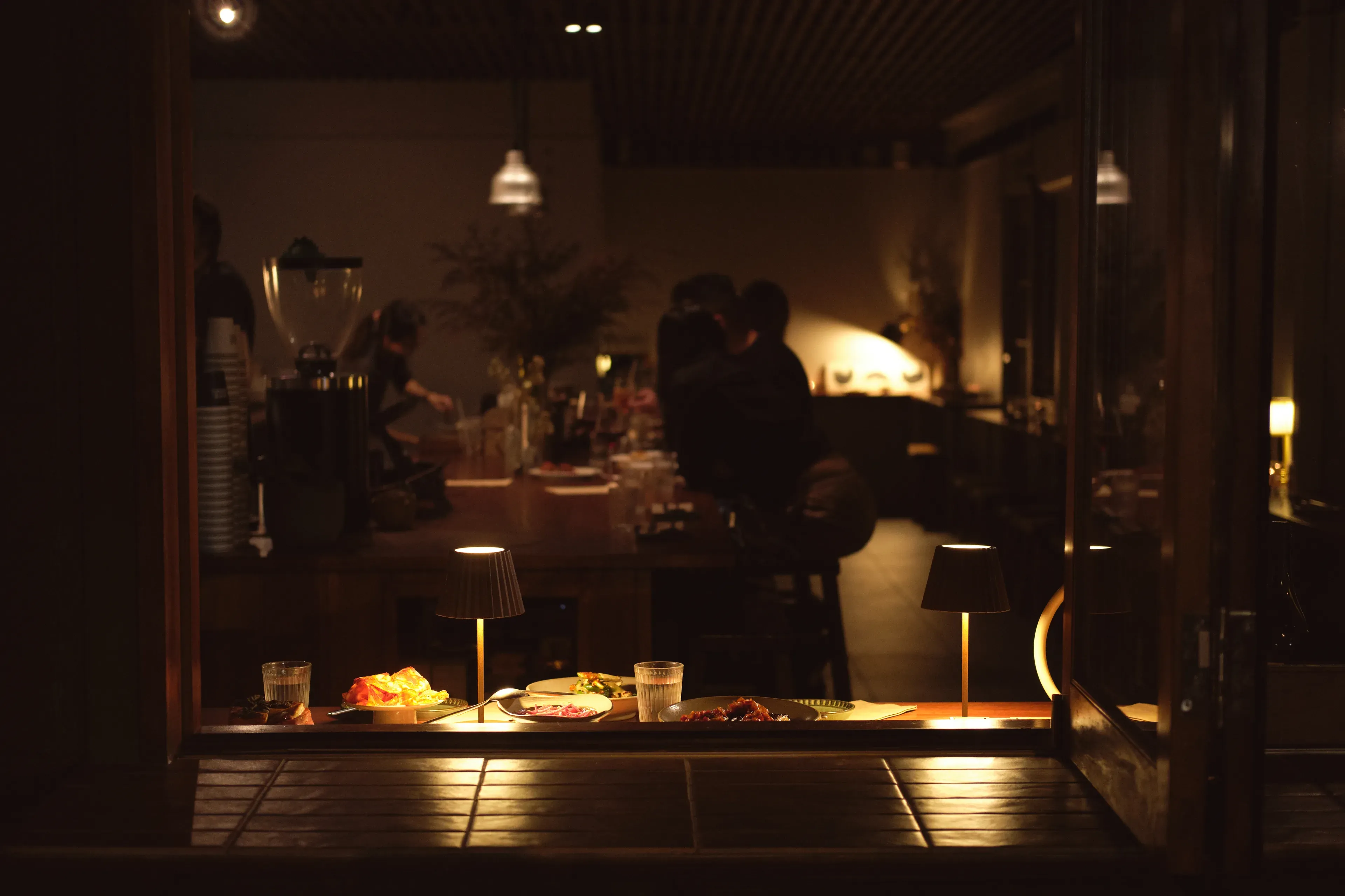 Snug at night through its open front window: plates, glasses and two small lamps on the sill, and behind them the timber bar, a cook at work and two diners seated